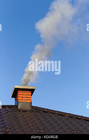 Fumo dalla ciminiera in mattoni sul tetto contro il cielo blu Foto Stock