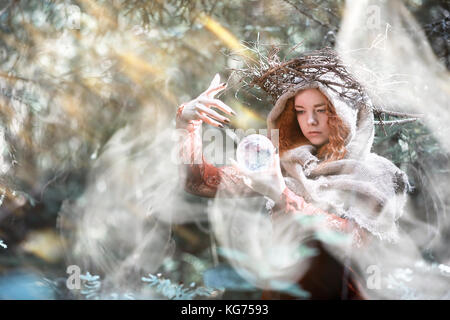 I capelli rossi strega in una fitta foresta con una sfera di cristallo e le erbe aromatiche Foto Stock