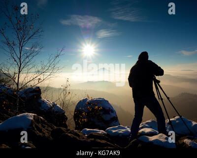 Fotografo prende foto di congelare autunnale di montagne, rocce ricoperte di polvere fresca neve. stony rock peak è aumentato da foggy valley. Foto Stock