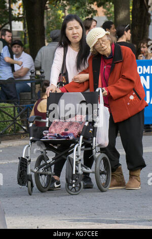 Un giovane americano asiatico accompagna la donna quale potrebbe essere la sua nonna per una passeggiata al di fuori della sua wheenchair. In Union Square Park a New York City. Foto Stock