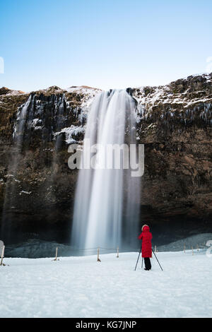 Cascata in inverno, fotografo in giacca rossa in piedi da sola sulla neve a cascata seljalandsfoss in Islanda Foto Stock