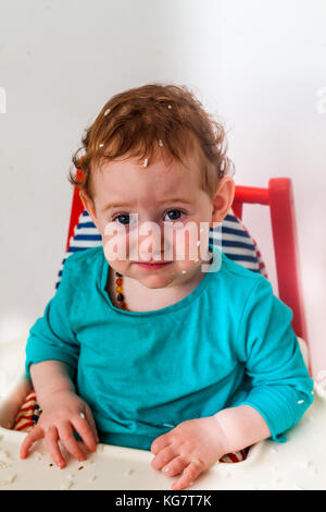 Confuso baby rendere divertente offeso in faccia il seggiolone con riso su tutta la testa - Concetto di sviluppo Foto Stock