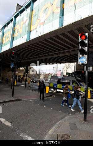 Persone di passaggio semaforo sotto il Camden Lock ponte mentre mangiare la pizza; polizia van in background. Foto Stock