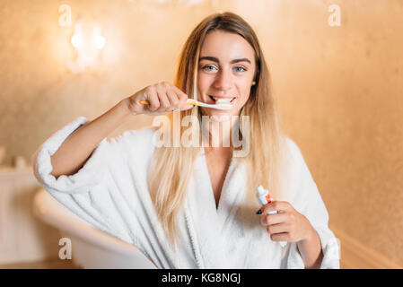 Giovane donna in accappatoio bianco spazzolare i suoi denti con spazzolino da denti, bagno interno su sfondo. mattina igiene della bocca, la pulizia dei denti Foto Stock