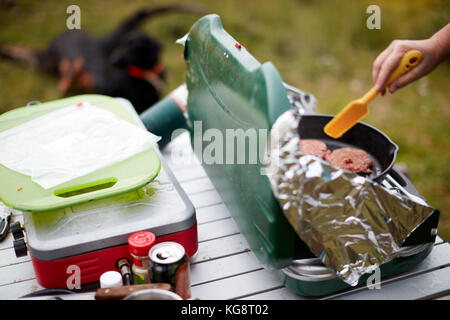 L'uomo la cottura di materie gli hamburger su un campeggio fornello a gas all'aperto su un piccolo tavolo portatile in una vista ravvicinata della sua mano, carne e la spatola Foto Stock