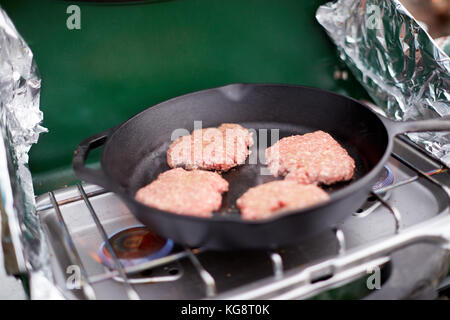 Materie di macinato di manzo gli hamburger in una padella su un campeggio bruciatore a gas con un foglio di alluminio protezioni del vento durante la cottura all'aperto Foto Stock