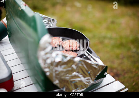 Cucinare gli hamburger su un bruciatore di gas all'esterno con un foglio di alluminio protezioni del vento impostato su un piccolo tavolo portatile mentre camping Foto Stock