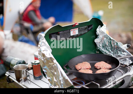 Polpette di carne macinata cottura su un bruciatore a gas a contatto con la natura in un campeggio con un uomo sfocata visibile in background sul campeggio Foto Stock