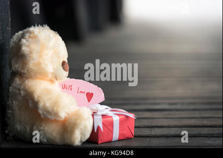 Gifting tema con un orsacchiotto di peluche toy tenendo un rosso avvolto confezione regalo e un pezzo di carta con il messaggio "con l'amore" Foto Stock