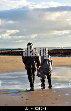 Un anziano o coppia di anziani camminare insieme sulla spiaggia in una giornata autunnale. anziane marito e moglie camminando su di una spiaggia di sabbia su breezy day con le nuvole Foto Stock