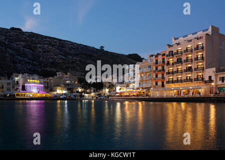 Il villaggio sul mare di Xlendi (pronunciato Shlendy) a Gozo, Malta, al tramonto, con lungomare alberghi e ristoranti si accende. Il turismo nel mediterraneo. Foto Stock