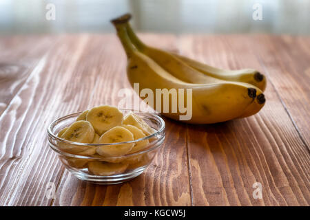 Fette di banana in un recipiente e tutta la banane sul tavolo di legno Foto Stock