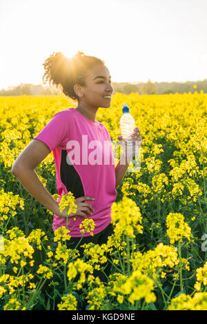 Outdoor ritratto della bella felice razza mista americano africano ragazza adolescente di sesso femminile donna giovane atleta runner acqua potabile da una bottiglia in un fiel Foto Stock