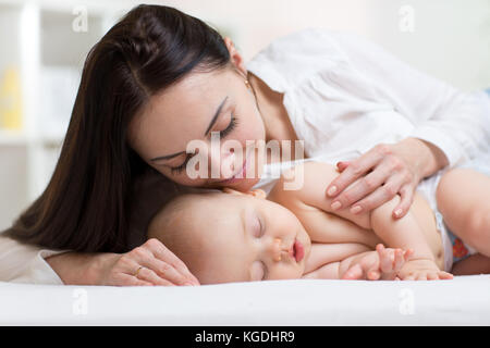 Bella giovane mamma guardando suo sonno bambino più piccolo e sorridente. Bambino giacente nel letto di casa Foto Stock