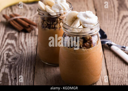 Zucca frullato di spezie con granola e banane conditi con panna montata su tavola in legno rustico Foto Stock