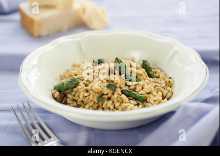 Risotto agli asparagi in vaso di ceramica sulla tovaglia viola con una forcella di argento e pezzi di formaggio parmigiano reggiano in background Foto Stock