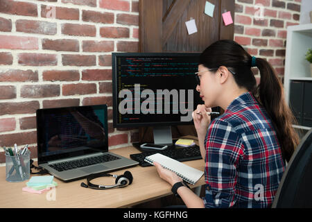 Giovane bella femmina di lavoratore di ufficio a lavorare sulla programmazione e la società holding notebook personale guardando mobile portatile pensando nuova idea progettuale. Foto Stock