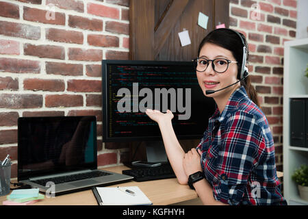 Fiducioso piuttosto azienda di programmazione del centro di servizio operatore femmina guardando la fotocamera e rendendo la postura di presentazione che mostra il codice della lingua. Foto Stock