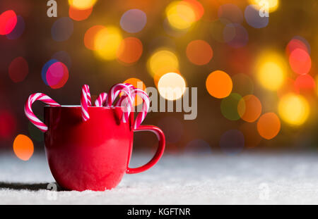 Coppa del rosso, rosso mug con la caramella canne in neve con defocussed luci fairy, bokeh in background, festa di Natale sfondo con copyspace Foto Stock