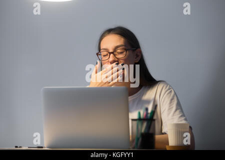 Stanco giovane femmina adulta segretario in vetri funziona su computer portatile al lavoro escursioni a tarda notte, sbadiglio, si copre la bocca con la mano. Foto Stock