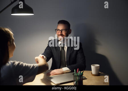 Due imprenditori di sigillare un affare con handshake dopo lunghe trattative in ufficio scuro con lampada luce. sorridente uomo barbuto in bicchieri agitare le mani con Foto Stock