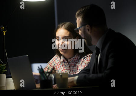 Due sorridenti colleghi impegnati sul progetto utilizzando il computer portatile o la negoziazione nel buio serale office, avente una buona volta, godendo di lavoro. enthusia Foto Stock