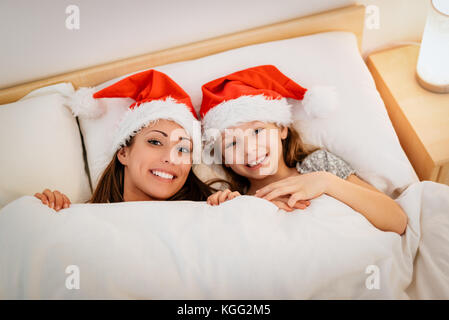 Giovane madre con sua figlia coperti con coperta e godere a letto alla vigilia di Natale, in attesa di santa claus. Foto Stock