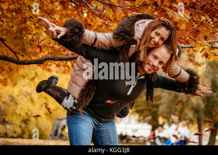 Bella coppia Giovane godendo un piggyback nel soleggiato parco in autunno colori. guardando la fotocamera. Foto Stock