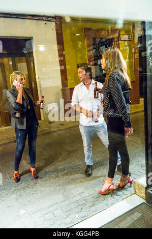 Due donne in tacchi alti e un uomo di bere birra e fumare in strada, Siviglia, in Andalusia, Spagna, visto attraverso la finestra della barra Foto Stock