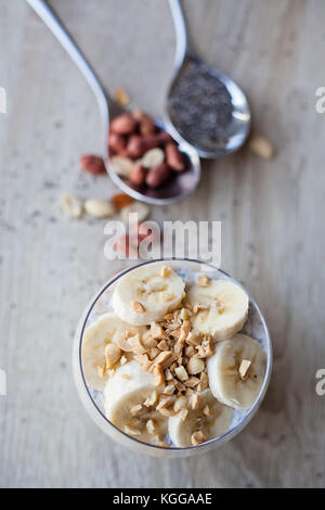 Banana chia budino di yogurt decorata con maturi fette di banana e le arachidi, cucchiai riempito con semi di Chia e dadi in background Foto Stock