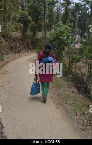 Donna nepalese con il bambino sulla schiena camminando su una strada sterrata in un piccolo villaggio, Panauti, nelle montagne himalaya del Nepal. Kameez tradizionale. Foto Stock