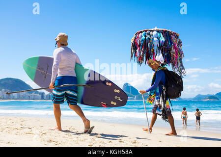 Rio de Janeiro - marzo 21, 2017: brasiliano venditore a vendere bikini cammina al fianco di un uomo che porta il suo stand up paddle tavola da surf lungo la spiaggia di Copacabana. Foto Stock