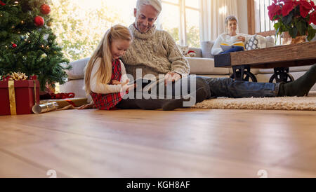 L'uomo anziano con il nipote seduto sul pavimento con tavoletta digitale. Bambina e grand i genitori nel soggiorno durante il periodo di Natale. Foto Stock