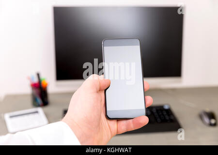 Primo piano di un uomo con smartphone con uno schermo vuoto monitor nelle mani, lavorando dietro la tastiera di un computer su un tavolo di legno in ufficio Foto Stock