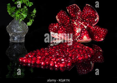 Aperto mature melograno con semi riflessa sul nero. piccolo riflesso vaso in vetro verde con foglie di prezzemolo in background. Foto Stock