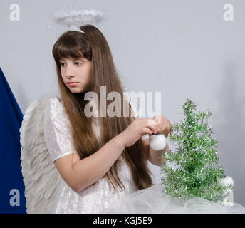 Una ragazza in un costume di angelo decora un piccolo albero di natale con le mele fatte di lana di cotone Foto Stock