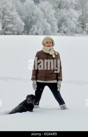 Una ragazza in una luce berretto lavorato a maglia e una scura giacca invernale è in piedi accanto a un grande nero labrador cane tra un campo coperto di neve. Foto Stock
