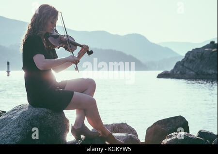 Donna retroilluminato suona il violino mentre è seduto sulla roccia sul lungolago contro le montagne Foto Stock
