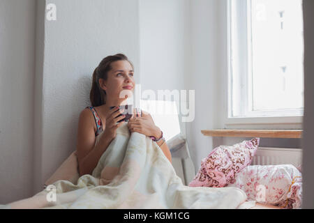 Considerato giovane donna seduta con tazza di caffè sul letto di casa Foto Stock