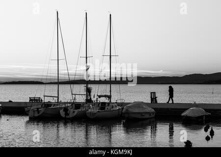 Un porto sul lago al tramonto, con varie barche e una donna a parlare al telefono mentre si cammina Foto Stock