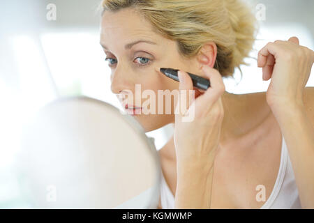 Di mezza età donna bionda mettendo il correttore oculare nella parte anteriore dello specchio Foto Stock