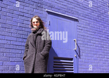 Hipster donna con occhiali da sole in piedi di fronte ad un luminoso viola un muro di mattoni. Indossare lana nero giacca e sciarpa colorata. Foto Stock