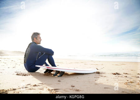 Surfer seduto sulla spiaggia sabbiosa con vista sull'oceano Foto Stock