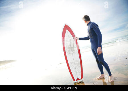 Surfer permanente sulla spiaggia sabbiosa, accanto alla tavola da surf Foto Stock