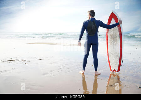 Surfer permanente sulla spiaggia sabbiosa, accanto alla tavola da surf Foto Stock