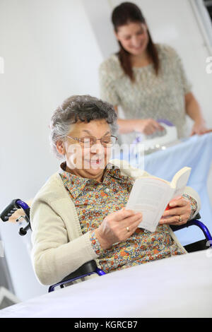 Donna anziana lettura libro mentre home helper ferri da stiro Servizio lavanderia Foto Stock