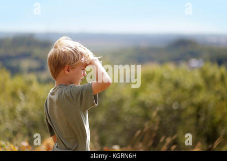 Un po' di 8 anno vecchio ragazzo su una escursione in natura, è schermatura gli occhi dal sole come egli si affaccia sopra gli alberi mentre in alto su un promontorio. Foto Stock