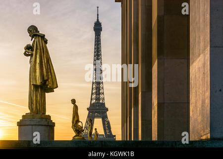 Il sole illumina il golden statue del Trocadero mentre la Torre Eiffel si staglia contro un cielo arancione. Foto Stock