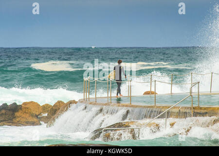 Un surfista maschio attende il momento giusto per entrare in acqua da Bronte Beach rock pool di Sydney, NSW, Australia in grandi condizioni surf Foto Stock