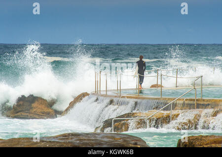 Un surfista maschio attende il momento giusto per entrare in acqua da Bronte Beach rock pool di Sydney, NSW, Australia in grandi condizioni surf Foto Stock